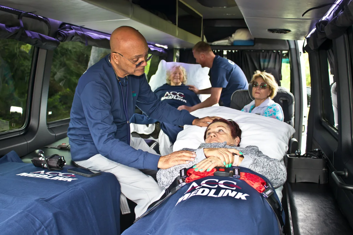 ACC Medlink medical attendant reassuring a patient in a bed, with a family member seated nearby