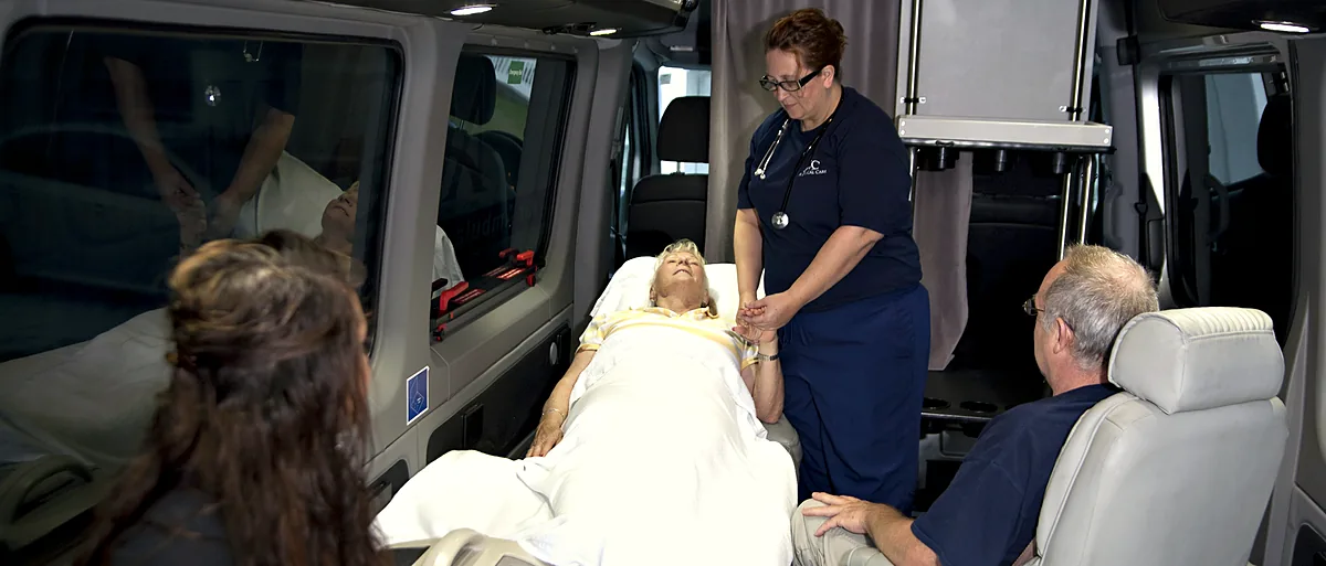 ACC Medlink attendant holding a patient's hand in bed while family members sit nearby