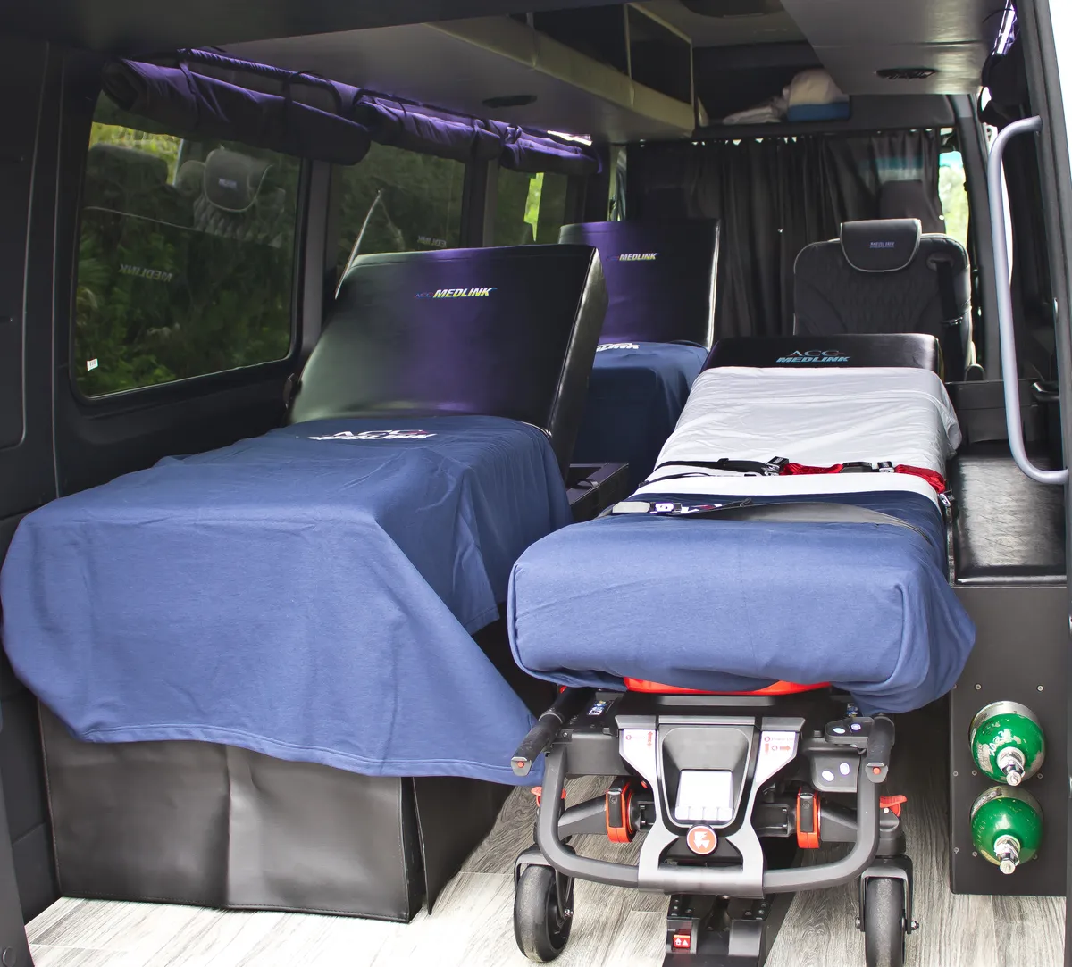Inside an ACC Medlink Sprinter: a power stretcher with a made-up mattress, oxygen cylinders on the wall, and a family seat up front
