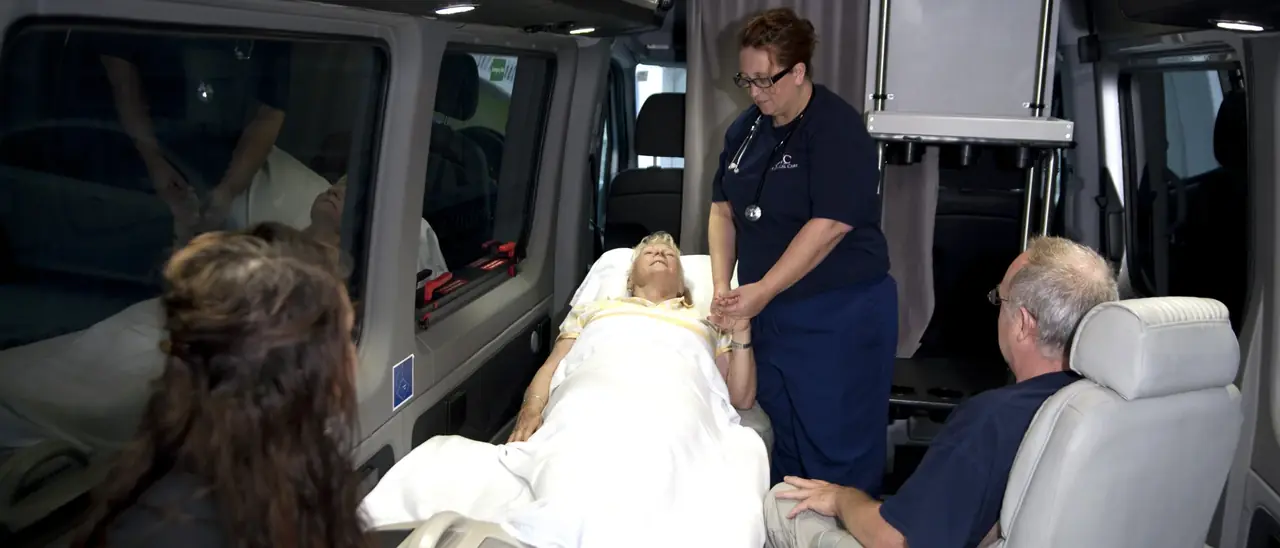 ACC Medlink attendant holding a patient's hand in bed while family members sit nearby