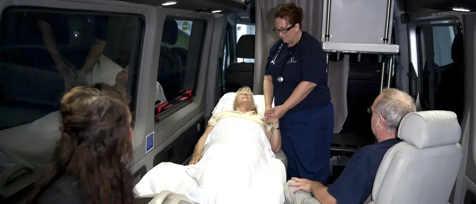 ACC Medlink attendant holding a patient's hand in bed while family members sit nearby